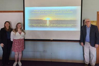 A student stands with two professors with a slide on a screen from their research